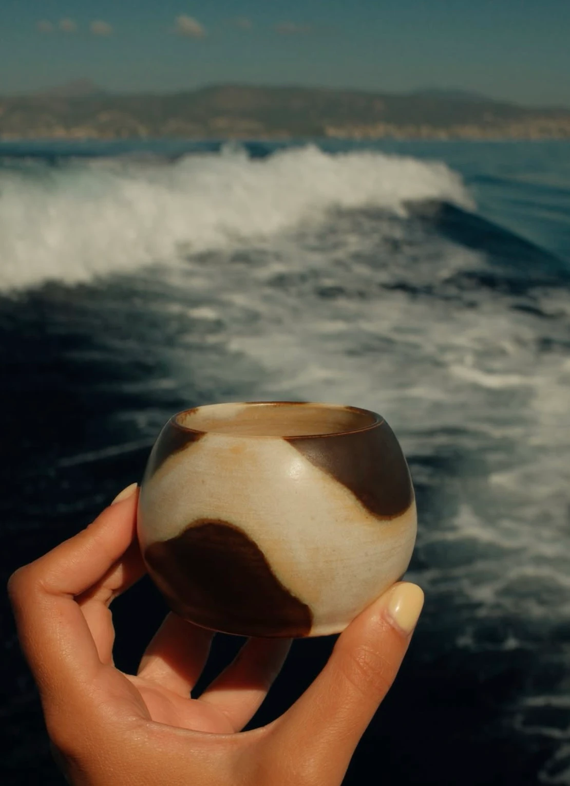 Clara’s cream and brown cup held in a hand beside the sea
